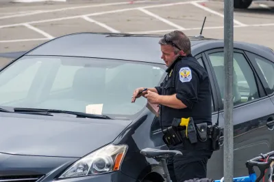 Male parking enforcement officer in uniform stands next to car with ticket in its windshield talking on phone or radio.