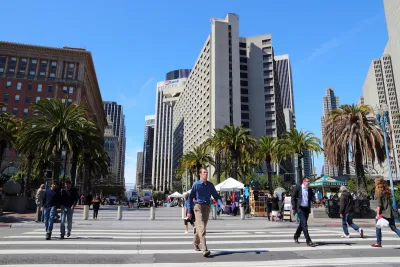People cross a wide street in San Francisco, with tall buildings in the background.