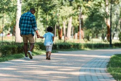 Dad and toddler holding hands walking down path in park.