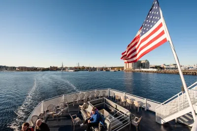 View from on board a Boston ferry with people sitting in front and an American flag flying from the boat on a sunny day.