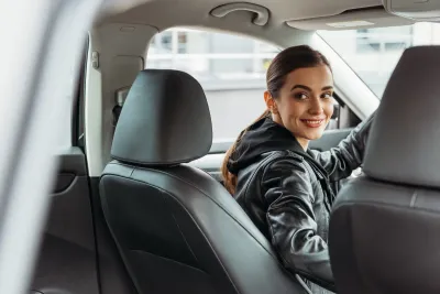 Young woman driver in black leather jacket looking back at passenger seat.