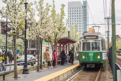 Green MBTA tram pulling up to station with people waiting under a flowering tree in Boston, Massachusetts.