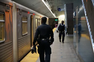 Security officer in dark blue uniform stands next to silver subway train on platform.