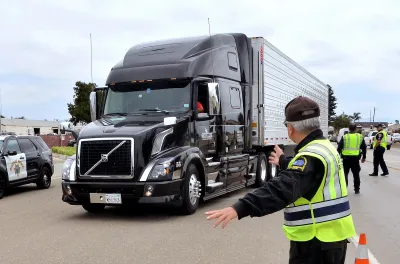 Black 18-wheeler truck being stopped by inspectors in black jackets and yellow safety vests.