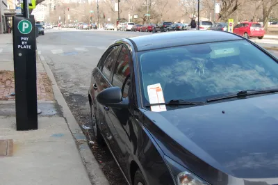 Dark-colored car parked at curb with white and red parking ticket in passenger side windshield wiper next to parking pay station with green P sign on sidewalk.