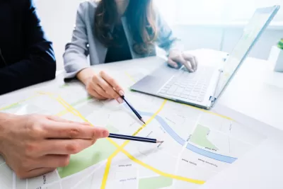 Close-up of two people pointing at a printed city map with pencils with a laptop in the background