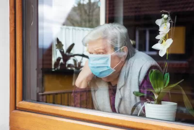 Elderly woman with short gray hair wearing blue surgical mask looks out window with wooden frame, a potted purple orchid on the windowsill