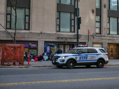 White NYPD SUV parked curbside next to unhoused people sitting on sidewalk.