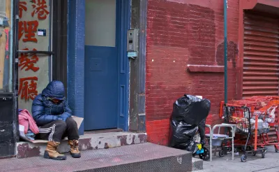 Unhoused person in blue parka sits huddled on a chair against a blue exterior door on red brick wall in New York City.