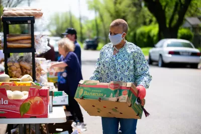 Food bank worker carries box of food in front of outdoor table filled with food items