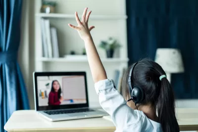 View from behind young girl student wearing headphones and raising her hand as she attends online class with dark-haired female teacher on laptop in front of her