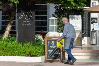 Man using lime green leaf blower in front of restaurant with sandwich board advertising mimosas in Miami Beach, Florida.