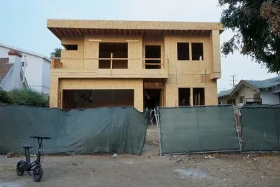 Boxy wood-frame two-story house under construction behind green construction fence in Los Angeles, California.