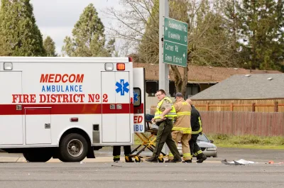 White ambulance and three first responders in yellow vests and firefighting suit wheel gurney into ambulance at car crash site at small intersection.
