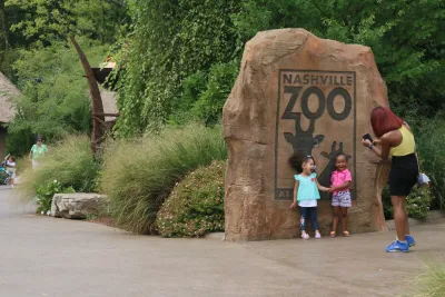 Two young children stand in front of Nashville Zoo logo on large rock as mother takes their photo.