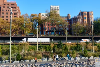 View of joggers on Brooklyn Bridge Park trail in foreground with Brooklyn-Queens Expressway traffic in background and brick buildings behind the highway