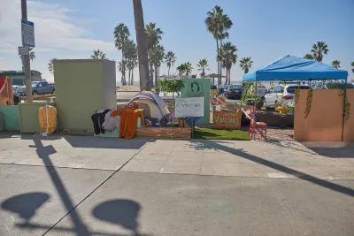Tents used by unhoused people on concrete walkway with Venice Beach, California beach and palm trees in background.