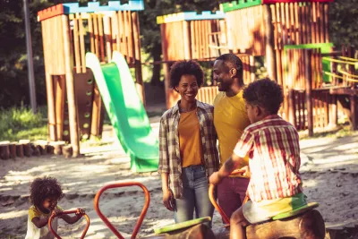 Two African American parents stand on playground with a young boy and toddler girl, looking happy on a sunny day.
