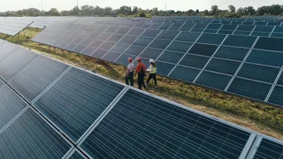 Three people in safety vests and hard hats walking through solar panels in open field.