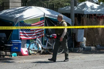 Sheriff's officer walks past tent with American flag draped on front in homeless encampment behind yellow police tape.