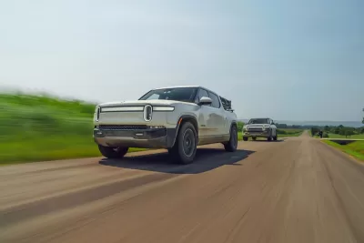 Two silver Rivian electric pick-up trucks driving down road with blurry grass on roadside