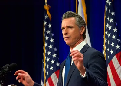 Gov. Gavin Newsom speaks with both hands half-raised. Two American flags, and the flag of California, are behind him.