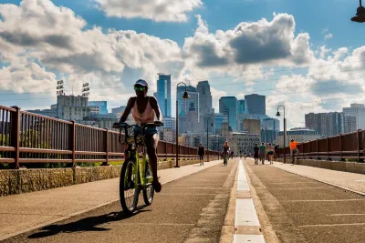 Woman rides bike on bridge in Minneapolis with downtown skyline in background on a sunny day.