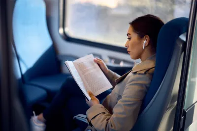 Woman reading book with white earbud in ear while sitting on train.