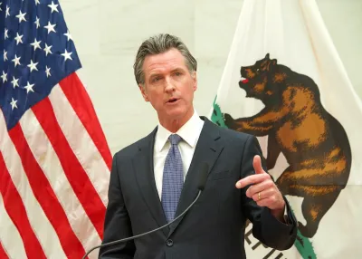 Gavin Newsom standing behind a podium, pointing at the camera. The flag of the United States and the flag of California are behind him.
