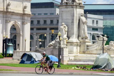 Cyclist rides past neoclassical buildings and homeless encampment in Washington, DC.