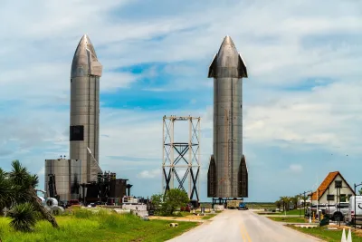 Two large rockets standing upright in Boca Chica, Texas behind Starbase sign with small older home in foreground.