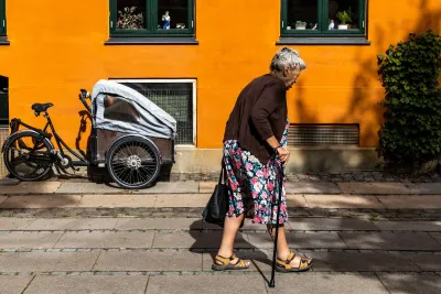 A senior woman with a cane walks by an old house with an ochre and orange color.