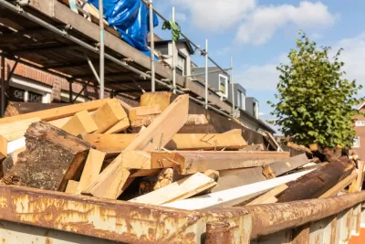 Old wood demolition waste, from destruction or renovation of buildings in a container on a work site in a street with a house. Wooden beams to be recycled into new building materials