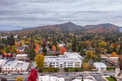 Aerial of Grants Pass, Oregon wth white city hall building in foreground and hills with colorfull fall leaves in background.