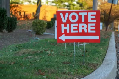 Red "Vote Here" sign with white text and arrow pointing left on green lawn next to curb