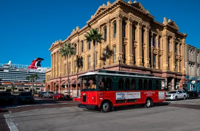 Historic turn of 20th century building in downtown Galveston, Texas with red streetcar passing by and large cruise ship visible in background.
