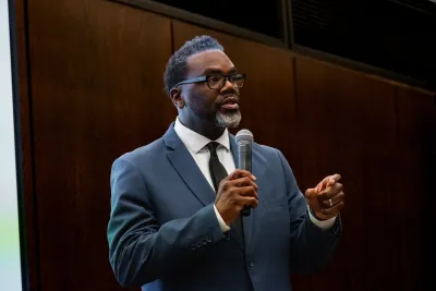 Chicago Mayor Brandon Johnson wearing dark suit speaking into a microphone in front of a dark wood wall.