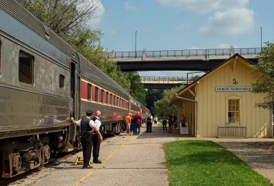 Passengers and crew stand outside of a train. there is a bridge ahead of them and a yellow A frame building.