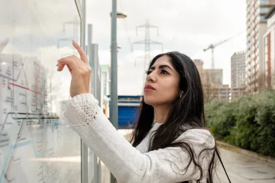 Young woman in white jacket looking at transit map outdoors with hand raised pointing to map.