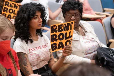 Young woman holding up "RENT ROLL BACK" sign in public meeting.