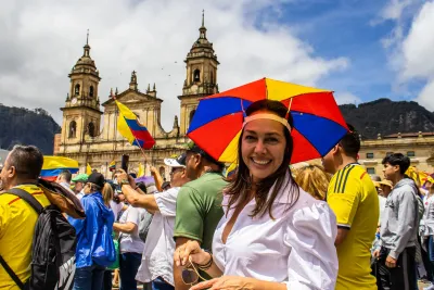 Woman wearing red, yellow, and blue umbrella hat smiling at camera during political protest in front of old Spanish-style yellow church in Bogota, Colombia.