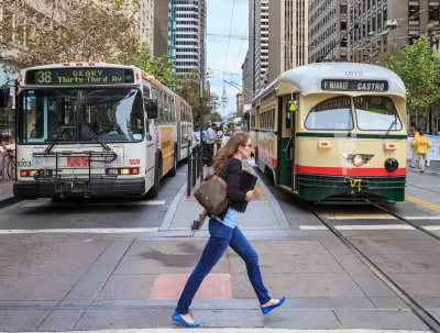 Woman in jeans and blue flats walks across a street in San Francisco with a vintage streetcar and a bus behind her.