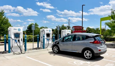 Small silver electric car plugged into white charger in parking lot in outdoor lot.