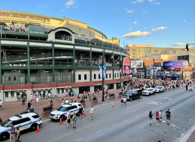 Wrigley Field, Chicago with two white police SUVs parked on street outside with people walking around.