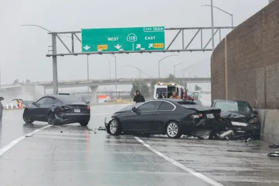 Three black cars crashed into each other on a rainy freeway under green exist signs with police officers standing next to cars.