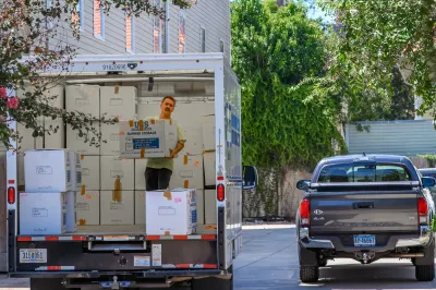 Man standing in moving truck stacking boxes on street with grey truck driving away.