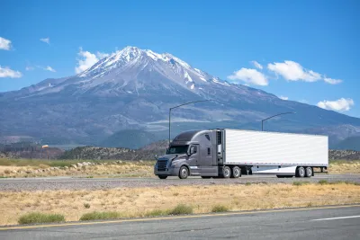 Grey big rig truck with white trailer driving on California highway with snow-capped peak visible in packground against blue sky.
