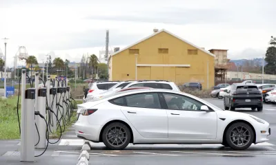White electric car charging at a station in a parking lot with a yellow barn in background.