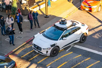 White Waymo self-driving car at intersection in San Francisco with pedestrians taking photos from sidewalk.