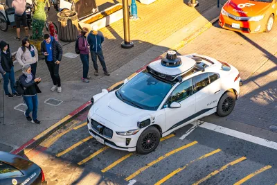 White Waymo self-driving car at intersection in San Francisco with people looking on from sidewalk.
